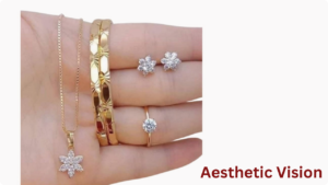 A hand displaying a matching minimalistic gold jewelry set with cubic zirconia details, including a fine pendant necklace, two textured bangles, flower stud earrings, and a solitaire ring. Maroon text in the corner reads "Aesthetic Vision."